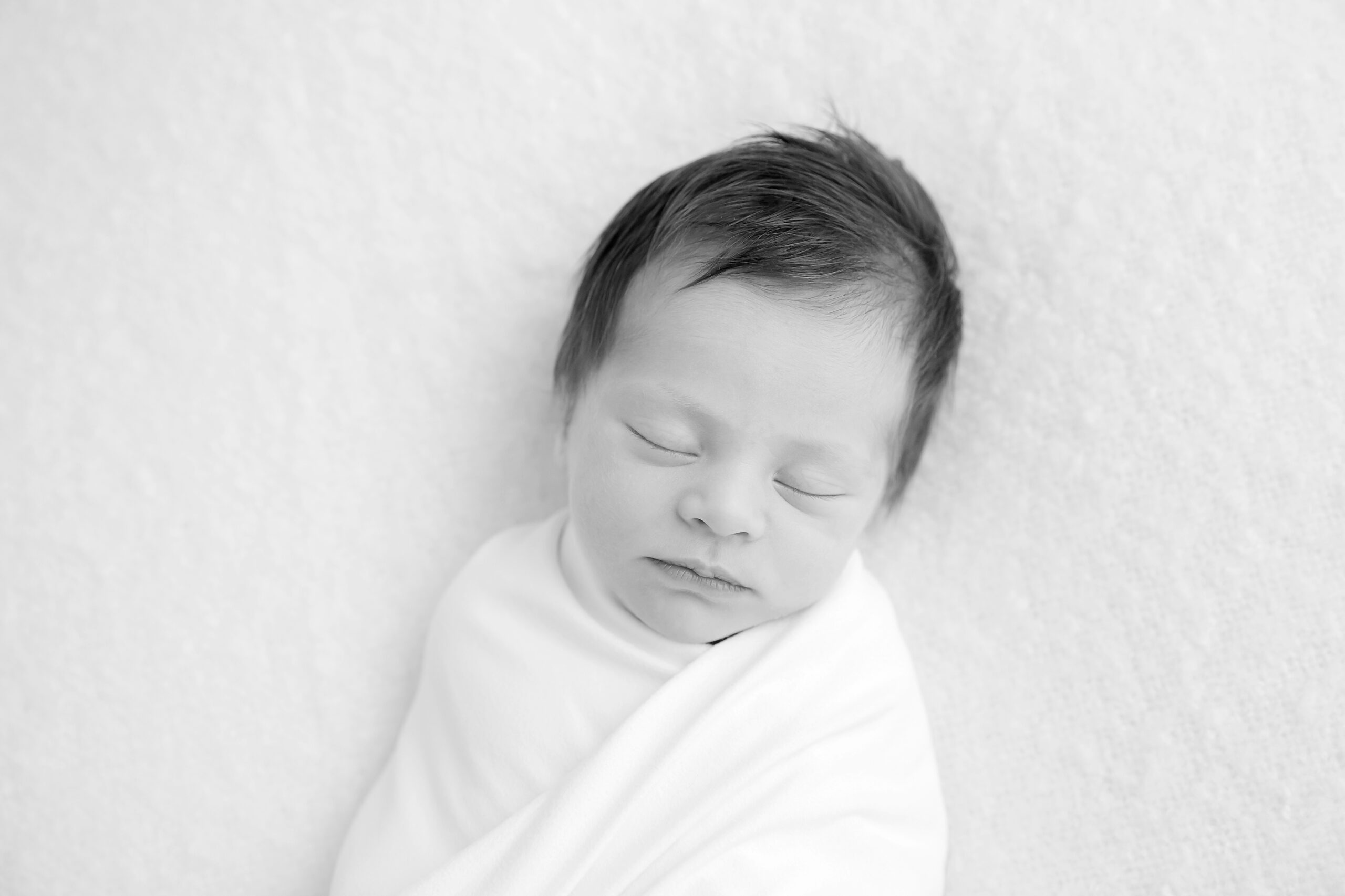 Black and white photo of a newborn baby girl with a head of dark hair, wrapped in a white swaddle