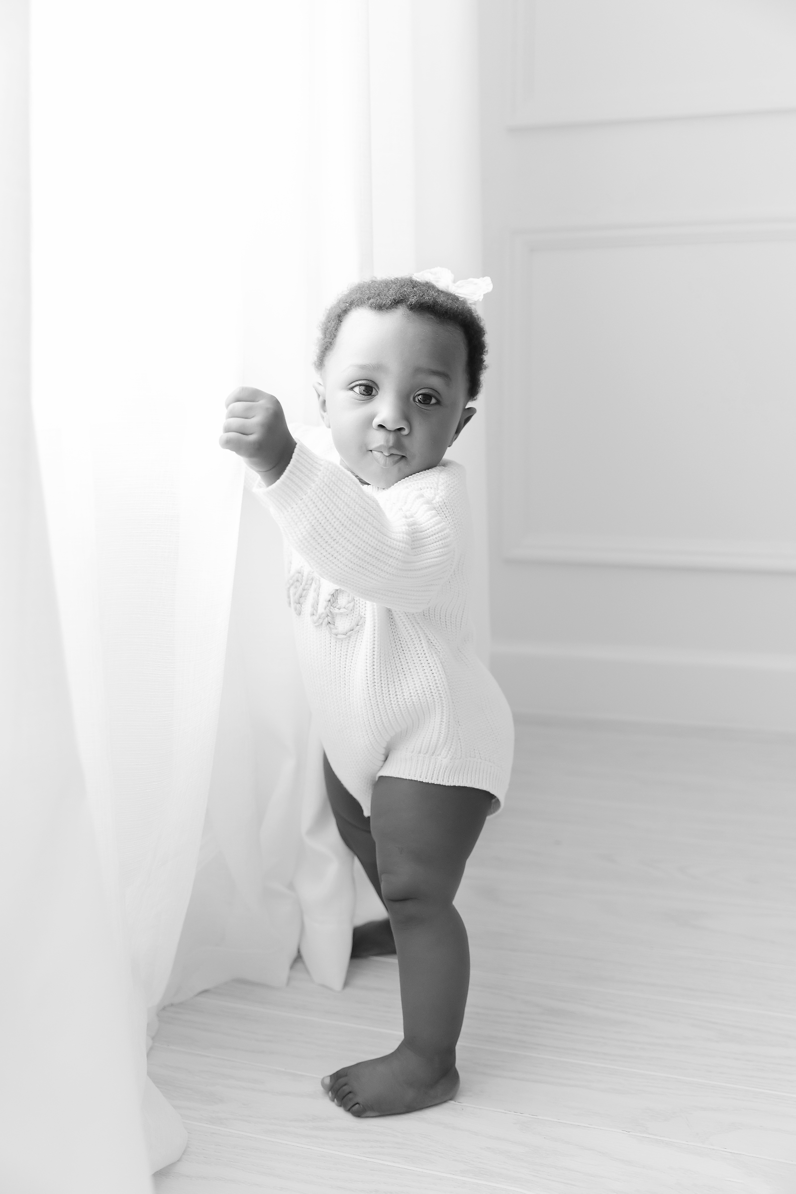 One year old girl stands at the window, holding onto the curtains