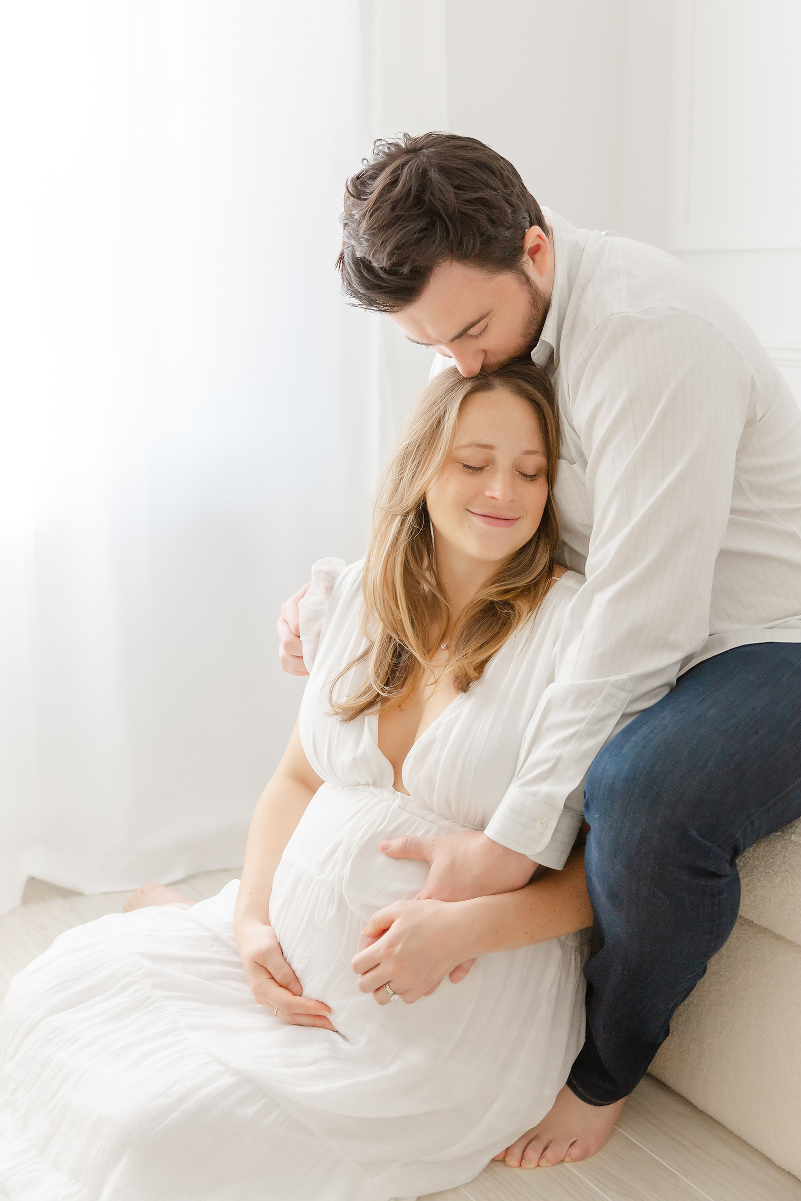 Pregnant woman seated on the floor entertwined in the arms of her husband who sits behind her