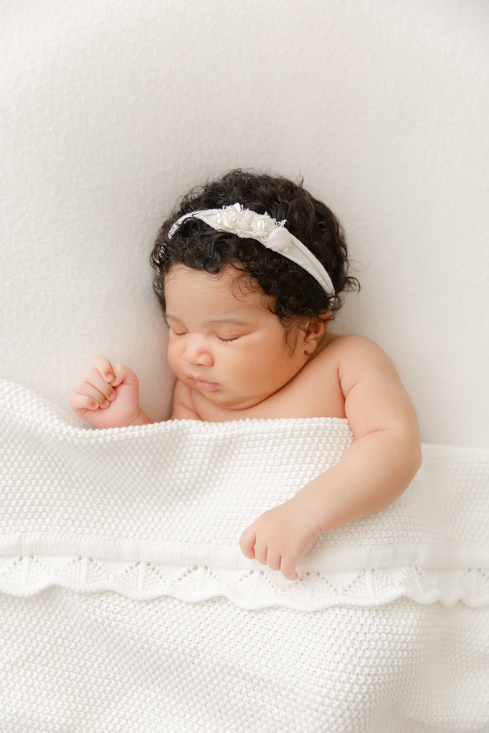 Newborn baby girl lays with a white headband and a scalloped white knit blanket