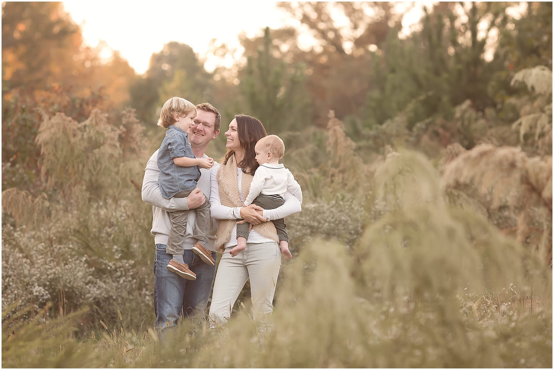 Family of four wearing a mix of denim and neutral outfits smile together in a field during their family photo session with Christy Johnson Photography