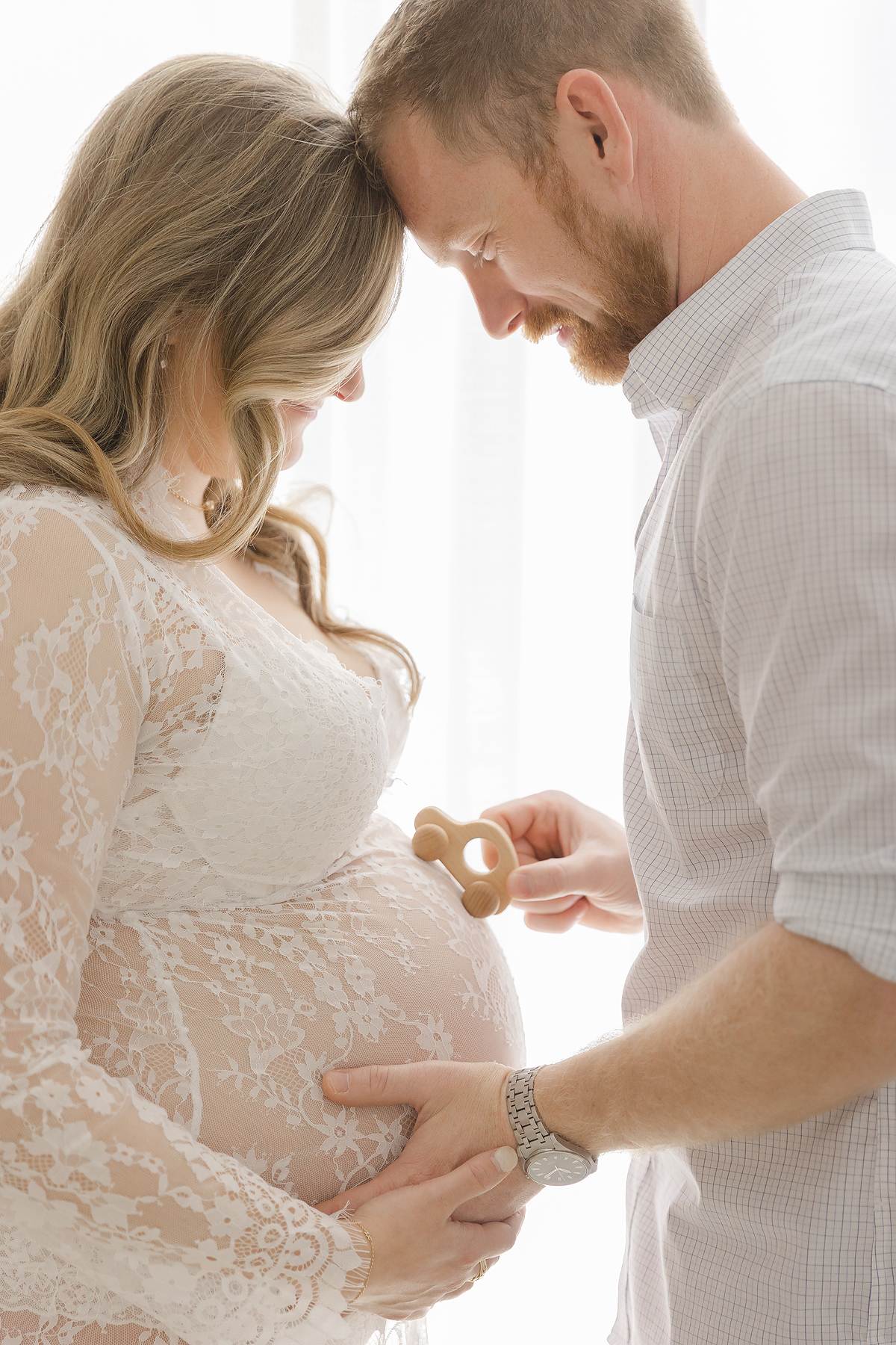 Pregnant woman and her partner cuddle and play with a wooden car on her belly during their maternity portrait session at Christy Johnson Photography in Wake Forest NC